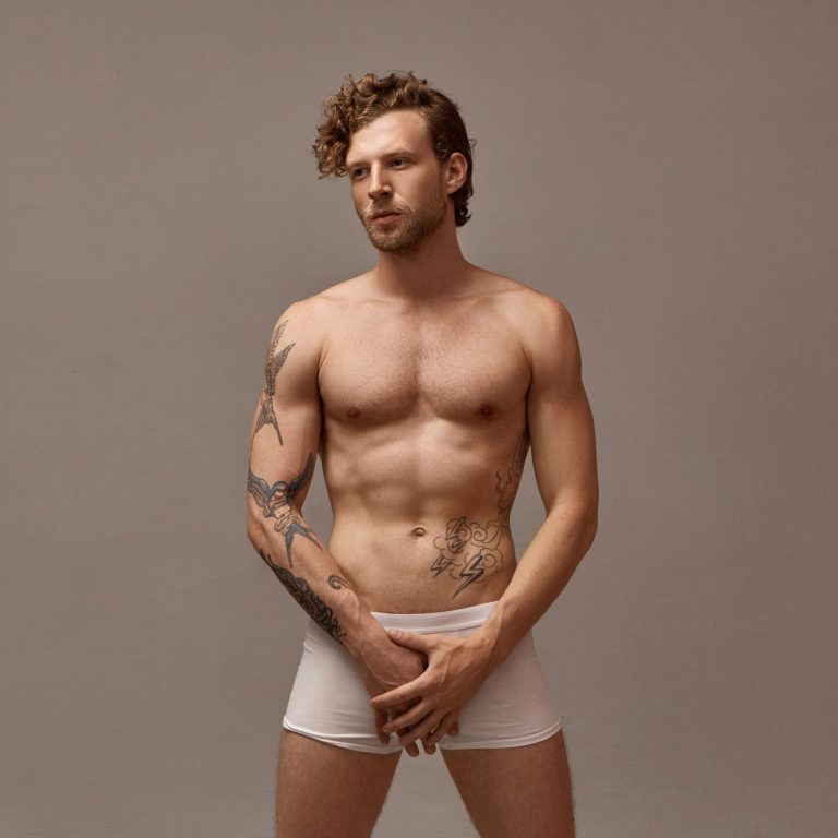 Isolated image of gorgeous handsome man with stubble and curly hairstyle posing naked in studio wearing only white boxer shorts, holding both hands on groin area, having serious facial expression